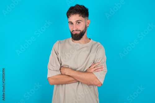 Self confident serious calm bearded caucasian man wearing casual T-shirt over blue background stands with arms folded. Shows professional vibe stands in assertive pose. © Jihan