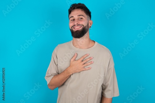 Joyful bearded caucasian man wearing casual T-shirt over blue background expresses positive emotions recalls something funny keeps hand on chest and giggles happily.