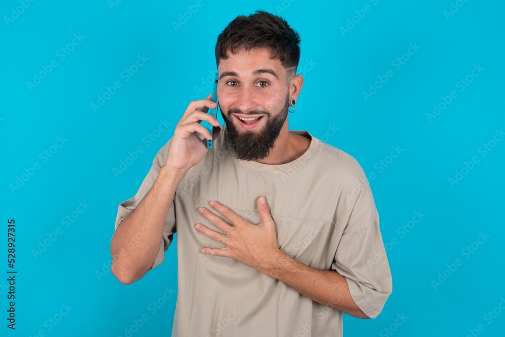 Smiling bearded caucasian man wearing casual T-shirt over blue background talks via cellphone, enjoys pleasant great conversation. People, technology, communication concept Smiling bearded caucasian man wearing casual T-shirt over blue background talks via cellphone, enjoys pleasant great conversation. People, technology, communication concept
