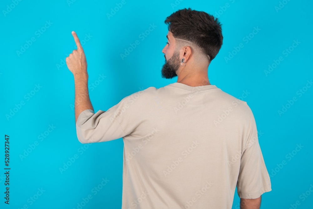 bearded caucasian man wearing casual T-shirt over blue background pointing to object on copy space, rear view. Turn your back bearded caucasian man wearing casual T-shirt over blue background pointing to object on copy space, rear view. Turn your back