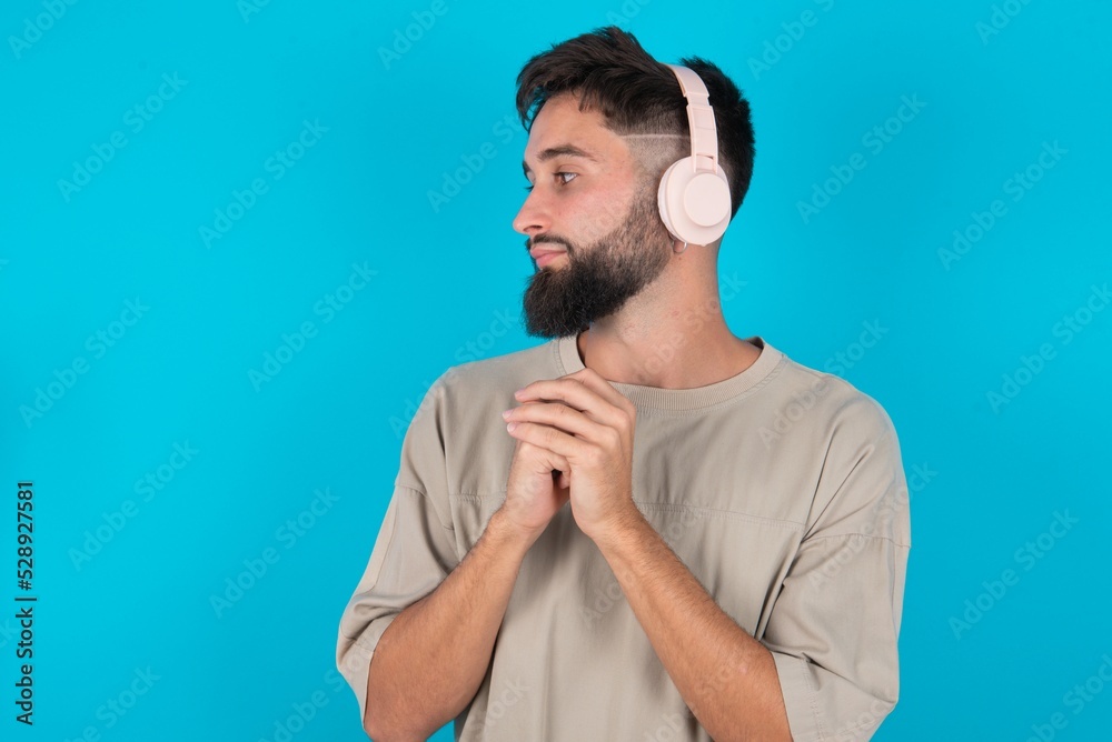 bearded caucasian man wearing casual T-shirt over blue background wears stereo headphones listening to music concentrated and looking aside with interest. bearded caucasian man wearing casual T-shirt over blue background wears stereo headphones listening to music concentrated and looking aside with interest.
