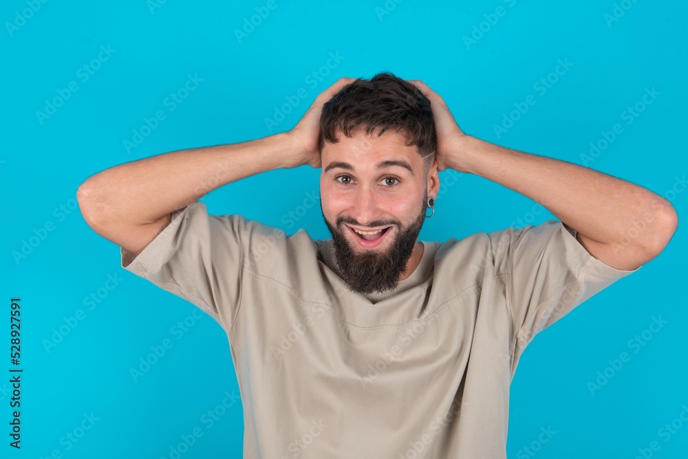 Cheerful overjoyed bearded caucasian man wearing casual T-shirt over blue background reacts rising hands over head after receiving great news. Cheerful overjoyed bearded caucasian man wearing casual T-shirt over blue background reacts rising hands over head after receiving great news.