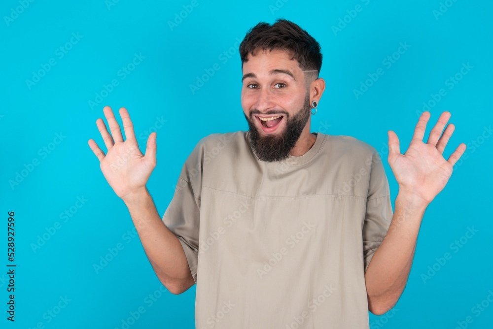 Delighted positive bearded caucasian man wearing casual T-shirt over blue background opens mouth and arms palms up after having great result Delighted positive bearded caucasian man wearing casual T-shirt over blue background opens mouth and arms palms up after having great result