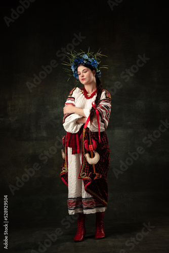 Art portrait of beautiful woman wearing traditional folk Ukrainian costume posing isolated over dark vintage background. Fashion, beauty, cultural heritage