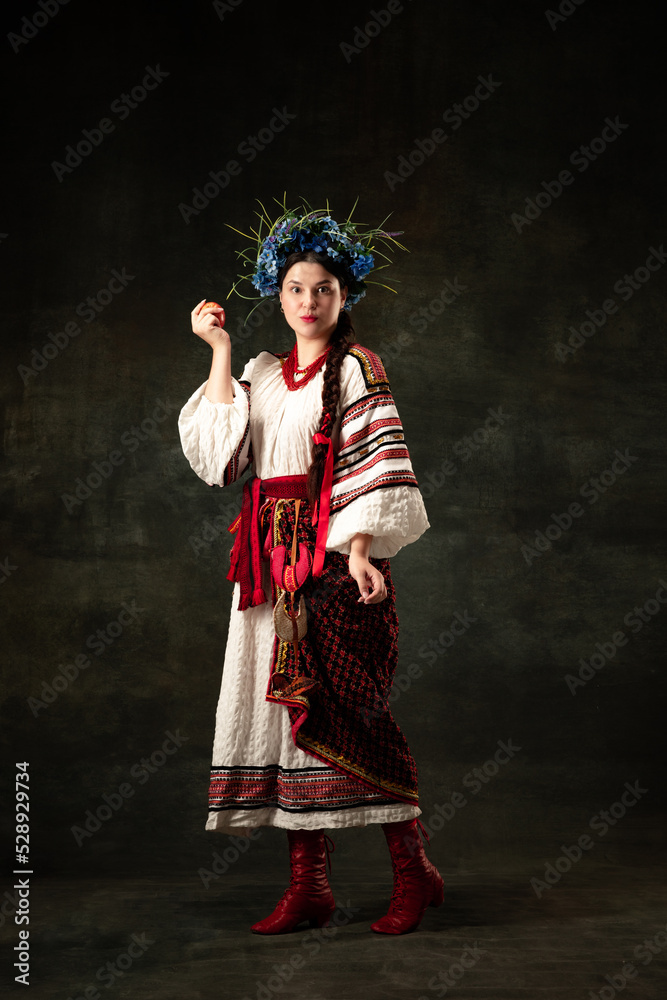 Adorable woman wearing national folk Ukrainian attire posing isolated over dark vintage background. Fashion, beauty, cultural heritage, eras comparison