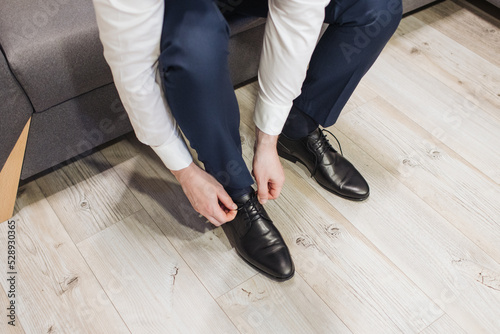 Black leather shoe lacing. Businessman in white shirt and suit trousers. Groom getting ready for the wedding. Wearing clothes background. Dressing up male fashion. Business fashion in hotel room.