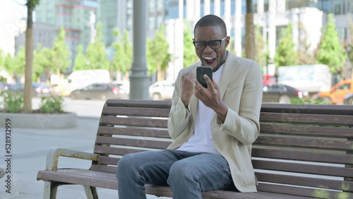 African Man Celebrating Online Success on Smartphone while Sitting Outdoor on Bench