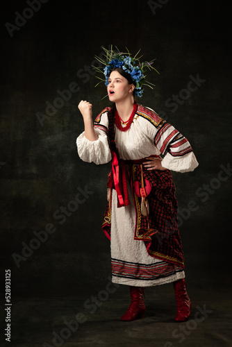 Young woman wearing national folk Ukrainian attire showing fist isolated over dark vintage background. Fashion, beauty, cultural heritage, eras comparison