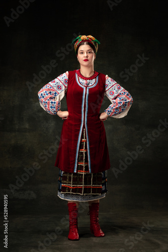 Art portrait of beautiful woman wearing traditional folk Ukrainian costume posing isolated over dark vintage background. Fashion, beauty, cultural heritage