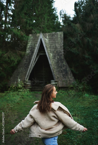 Rear view beautiful stylish model posing or dancing by vintage wooden hut chalet during travel in Carpathian dense forest photo