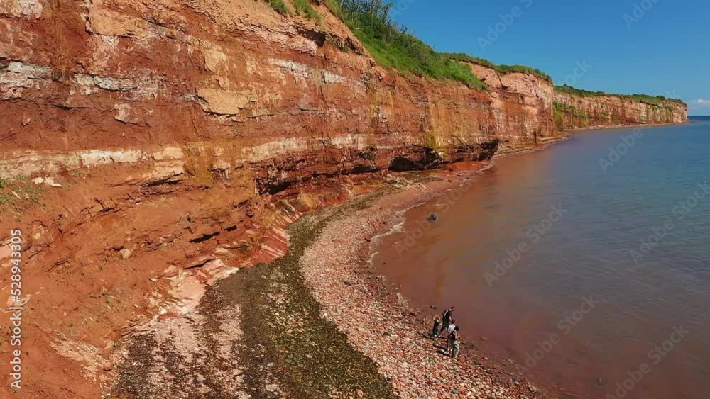 Aerial view with a drone of the red earth mountain of Caplan, rotating to the right and revealing a family playing on the edge of the Atlantic Ocean, in Gaspesie