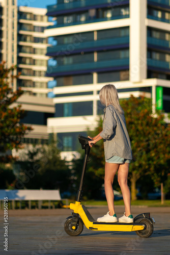 Young beautiful slender woman in denim shorts, shirt, riding an electric scooter in the park in summer, photo from the back.Modern girl,new generation, electric transport, ecological transport, sunset
