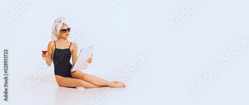 Portrait of beautiful young girl in black swimming suit relaxing, reading magazine, posing over grey background. Flyer