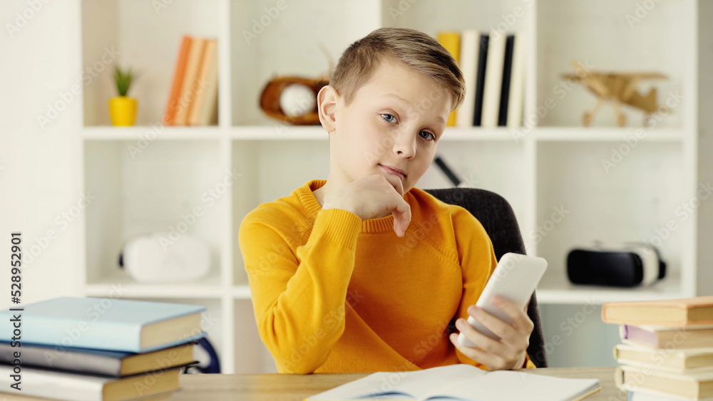 pensive schoolboy holding mobile phone near copy book and books on desk. pensive schoolboy holding mobile phone near copy book and books on desk.