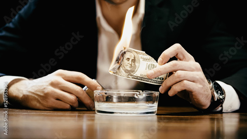Cropped view of businessman holding burning money and lighter near ashtray isolated on black.