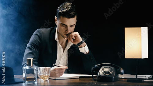 Businessman looking at notebook near telephone and whiskey on table on black background with smoke.
