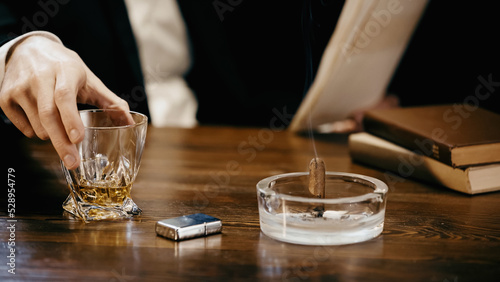 Cropped view of businessman holding glass of whiskey near cigar with smoke in ashtray isolated on black.