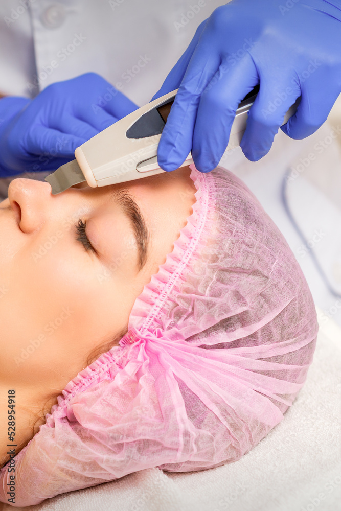 Fototapeta premium Young caucasian woman receiving facial skin cleaning by ultrasonic cosmetology face equipment in a medical salon