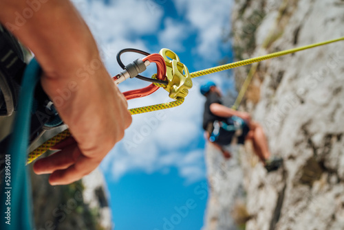 Belay device close-up shot with a boy on the cliff climbing wall. He hanging on a rope in a climbing harness and his partner belaying him on the ground. Active people and sports concept image photo