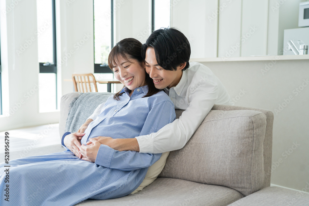 自宅で笑顔でお腹をさする日本人妻と後ろからハグをする夫