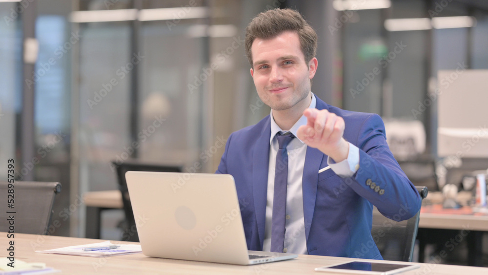Businessman Pointing at Camera while using Laptop in Office