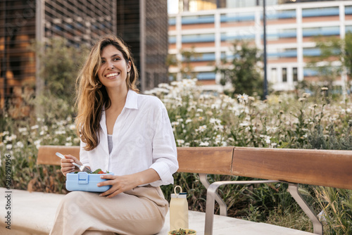 Foto Smiling young caucasian business woman is sitting on bench outside and eating salad
