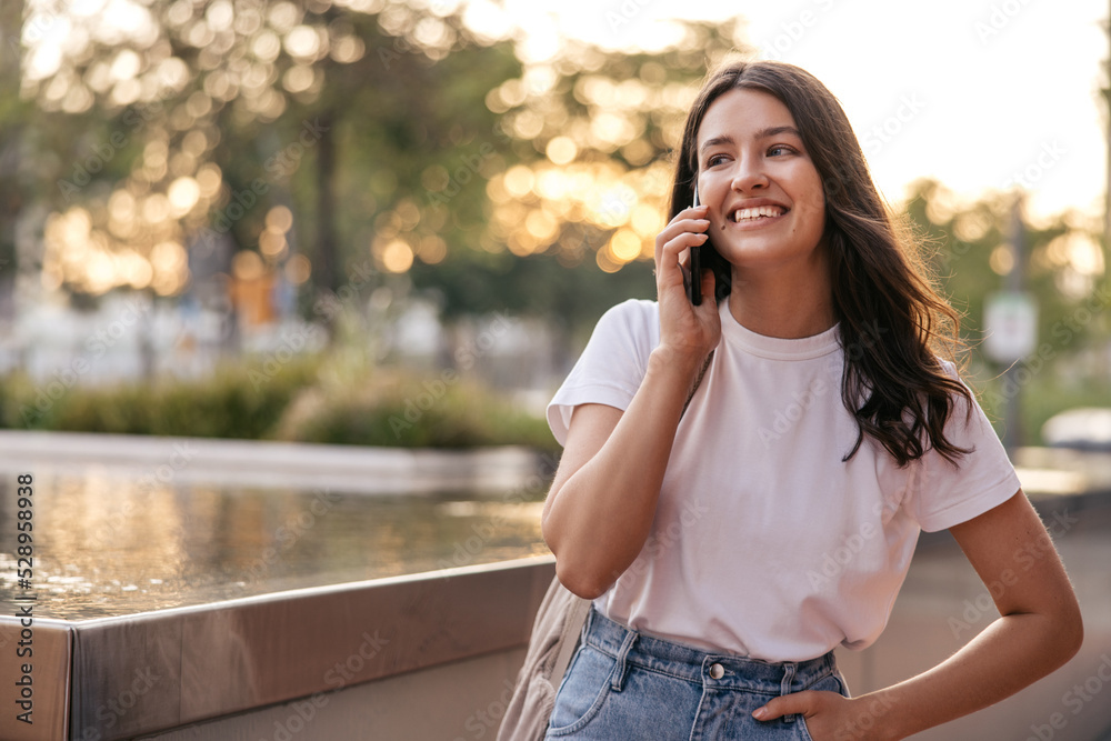 Cheerful young caucasian girl makes call using phone spending time in park summer. Brunette woman wears casual clothes. Concept lifestyle, technology Cheerful young caucasian girl makes call using phone spending time in park summer. Brunette woman wears casual clothes. Concept lifestyle, technology