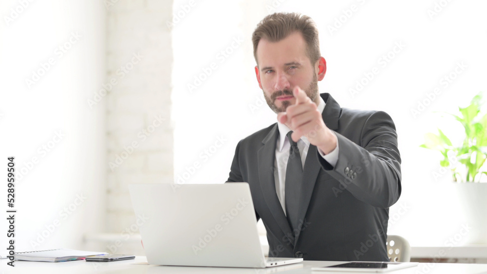 Young Businessman Pointing at Camera while using Laptop in Office Young Businessman Pointing at Camera while using Laptop in Office