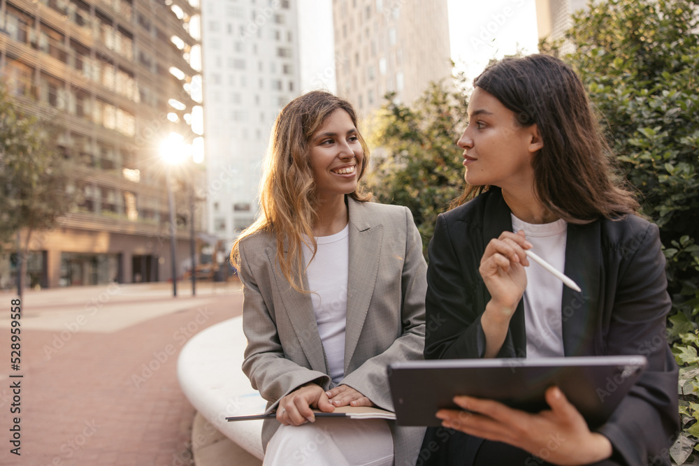 Successful young caucasian women standing near modern office holding tablet. Employees spending time outdoors on sunny day. Business people concept