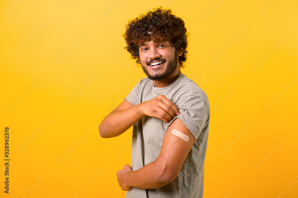 Smiling young curly Indian guy showing arm with band-aid after vaccine injection isolated on yellow background, vaccinated multiracial man feels safe with vaccine Smiling young curly Indian guy showing arm with band-aid after vaccine injection isolated on yellow background, vaccinated multiracial man feels safe with vaccine