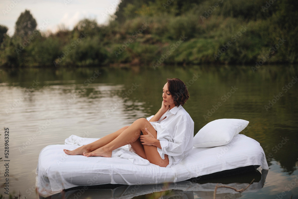 Brunette woman with short hair in a white shirt in bed on the water. A young woman floats on an inflatable mattress with a white sheet on the lake.