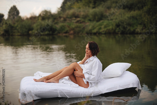 Brunette woman with short hair in a white shirt in bed on the water. A young woman floats on an inflatable mattress with a white sheet on the lake. © VikaNorm