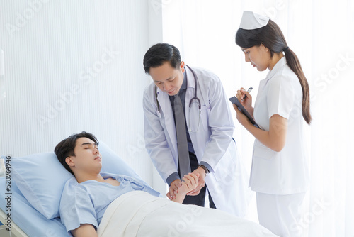 Senior doctor and young male patient who lie on the bed while checking symptom, consult and explain with nurse taking note and supporting in hospital wards.
