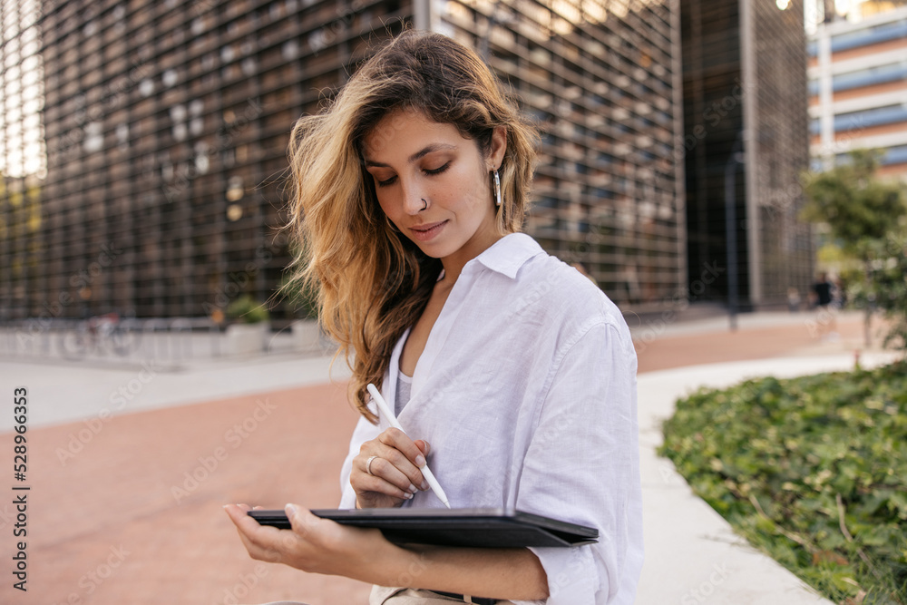 Working young caucasian woman with tablet sitting in office area outdoors. Blonde wears classic white shirt. Business finance concept Working young caucasian woman with tablet sitting in office area outdoors. Blonde wears classic white shirt. Business finance concept