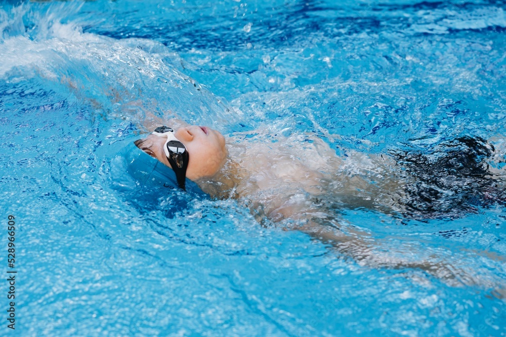 little caucasian boy wearing goggles swimming backstroke in a pool