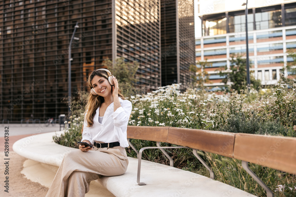 Pretty young caucasian woman with phone and headphones sits on bench against backdrop of buildings. Blonde wears shirt and pants in spring. Music concept.
