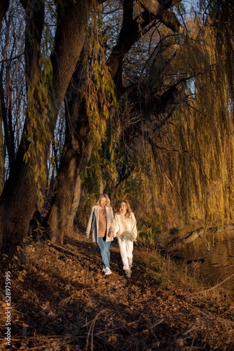 Adult daughter and her pregnant mom holding hands while standing in autumn park photo