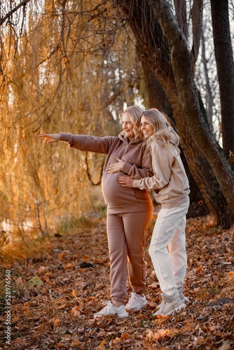 Adult daughter hugs her pregnant mom while standing in autumn park photo