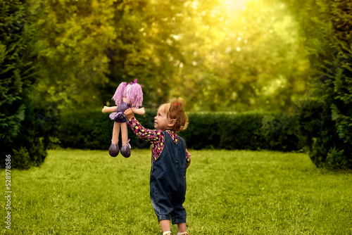 Cute little girl playing with her favorite doll outdoors in a summer park.