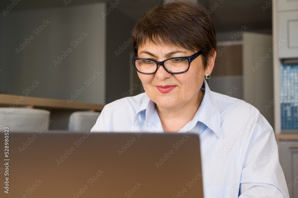 Closeup portrait of happy elder woman in glasses sitting in front of laptop and looking at the screen. Use of modern technologies by senior generation. Closeup portrait of happy elder woman in glasses sitting in front of laptop and looking at the screen. Use of modern technologies by senior generation.