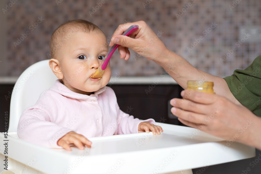 Baby food. A baby in a high chair eats vegetable puree from a spoon. Mom feeds the baby from a jar. Kitchen. Lifestyle. Baby food. A baby in a high chair eats vegetable puree from a spoon. Mom feeds the baby from a jar. Kitchen. Lifestyle.