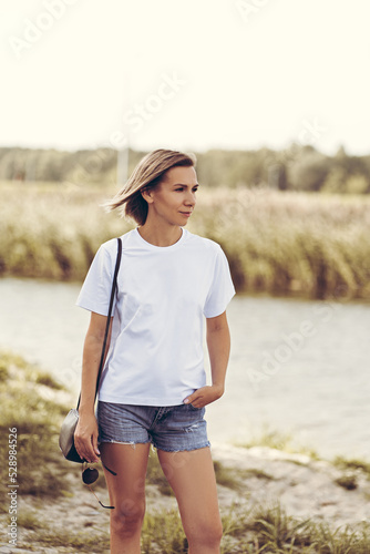A charming young woman in a white T-shirt and denim shorts walks along the river bank. Relax, walk, vacation © Olga