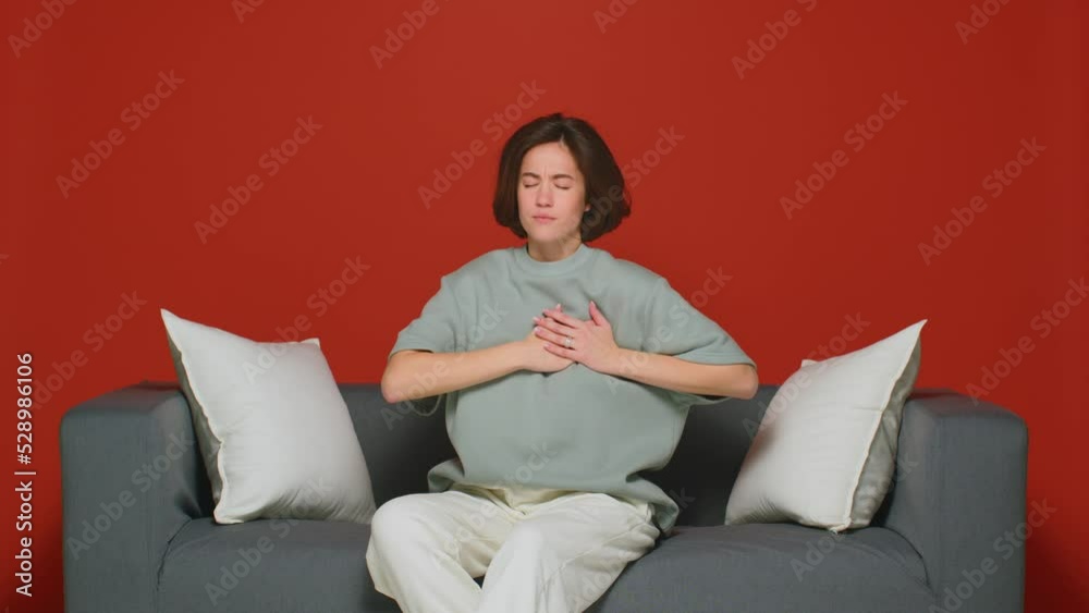 Ill young woman touching her breast, breathing suffering panic attack, heart decease, sitting on couch Ill young woman touching her breast, breathing suffering panic attack, heart decease, sitting on couch