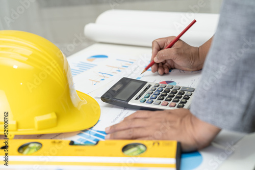 Asian engineer with architectural house plan project blueprint, graph and yellow helmet and engineering construction tools.