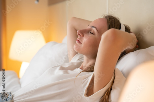 Young woman wearing white headphones listens to music. Brown-haired tourist relaxing on king-size bed enjoys spending vacation in luxury hotel resort