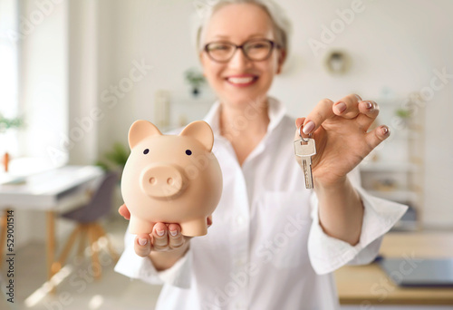 Fotografija Happy smiling middle aged woman holding a pink piggy bank and house keys