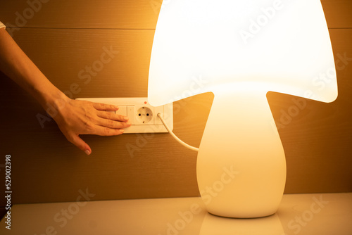 Hand of woman pushes button on white switch near socket to turn on lamp standing on bedside table. Bright light illuminates hotel room close view photo