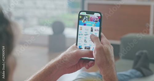 man is surfing the Internet on his cell phone at an online shopping and retail website. He is sitting on his comfortable sofa in his living room