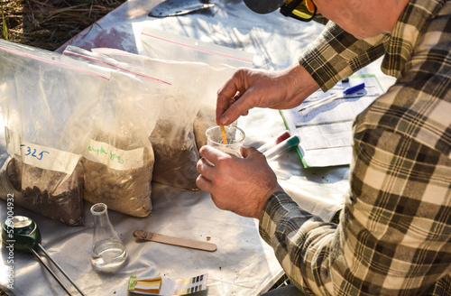 Male agronomy specialist testing soil sample outdoors, using laboratory equipment litmus paper, performing soil certification at agricultural grain field sunrise. Environment research