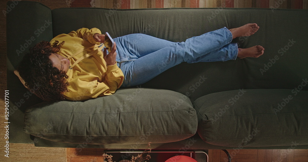 Young woman sits on the sofa in her living room and picks up her cell phone. One hispanic girl sending message and surfing the internet. From above angle of person browsing social media Young woman sits on the sofa in her living room and picks up her cell phone. One hispanic girl sending message and surfing the internet. From above angle of person browsing social media